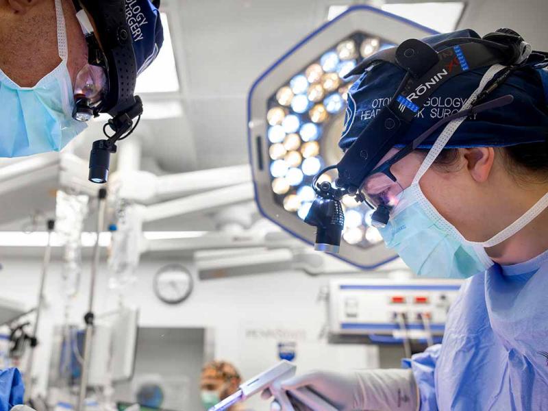 Two masked clinicians wearing surgical headlamps perform a procedure in an operating room under bright surgical lights.