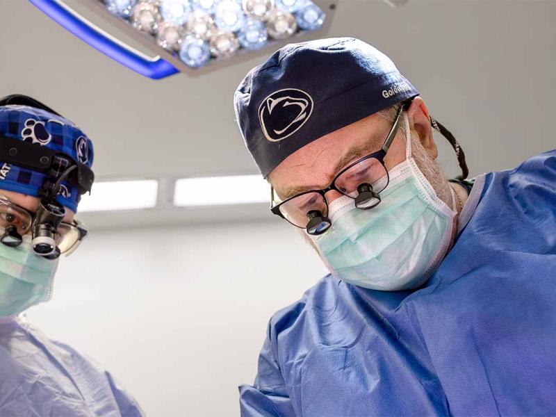Two Penn State Health surgeons, wearing masks and protective gowns performs a procedure in a modern operating room under overhead lights.