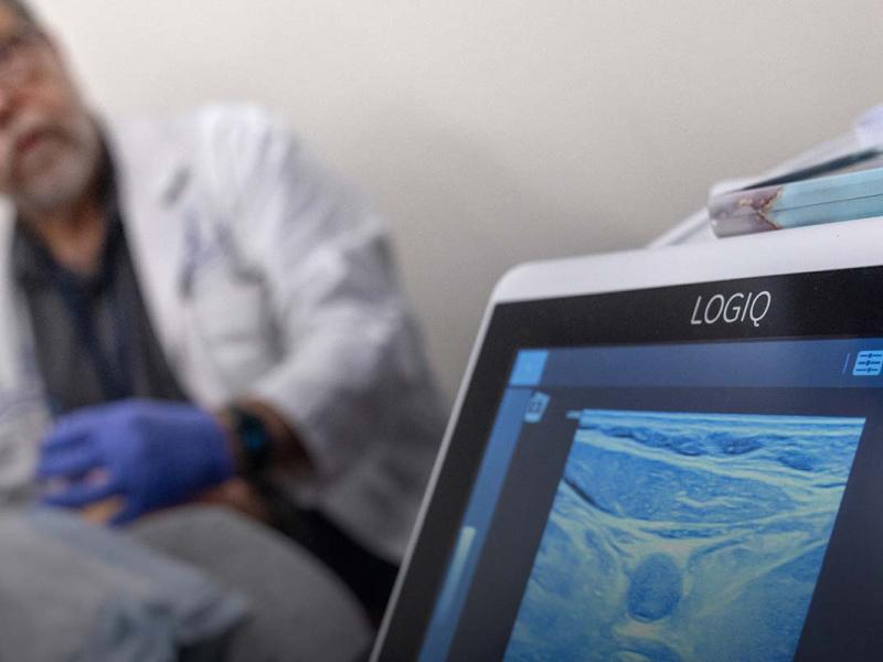 A closely cropped view of the ultrasound screen. In the background, Dr. David Goldenberg is seated using an ultrasound to guide a needle and microelectrode probe into the patient's affected area of thyroid.