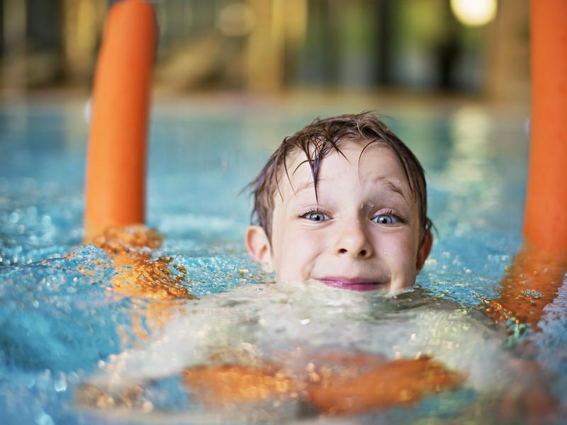Happy kid learning to swim in indoors swimming pool. The boy is aged 5  and is using orange pool noodle. He is smiling into the camera.