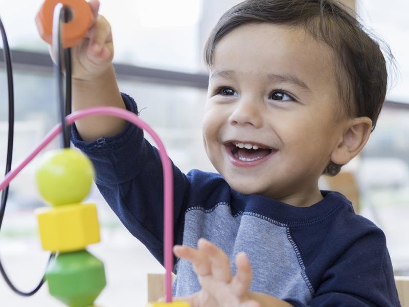 An adorable toddler boy sits at a table in a doctor's waiting room and  reaches up cheerfully to play with a toy bead maze.