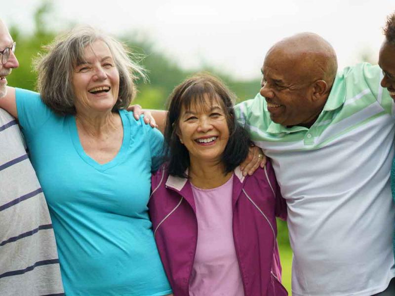 Five adults stand close together outdoors on a grassy area with trees in the background, each with an arm around the next person’s shoulders. Faces are turned slightly toward one another, smiling, conveying closeness and connection in natural daylight.