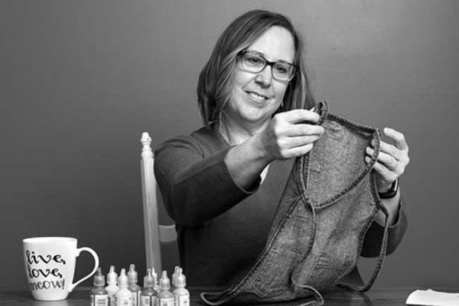 Wendy Johnson, who has shoulder-length hair and wears glasses, sits behind a table and smiles as she looks at a child’s apron she holds in front of her. Bottles of fabric paint and a mug that says “Live, Love, Meow” are on the table.