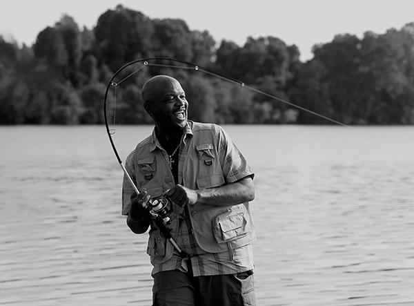 A man, dressed in a fishing vest, stands by a lake and smiles as he reels in his fishing line. His fishing rod is bent over his head.