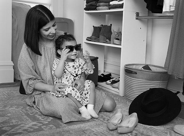 Alyssa Acevedo, who has shoulder-length hair and wears a long-sleeve dress, sits on her bedroom floor with her toddler daughter, who wears a print dress and sunglasses, on her lap. Shoes and clothes surround them on closet shelves and on the floor.